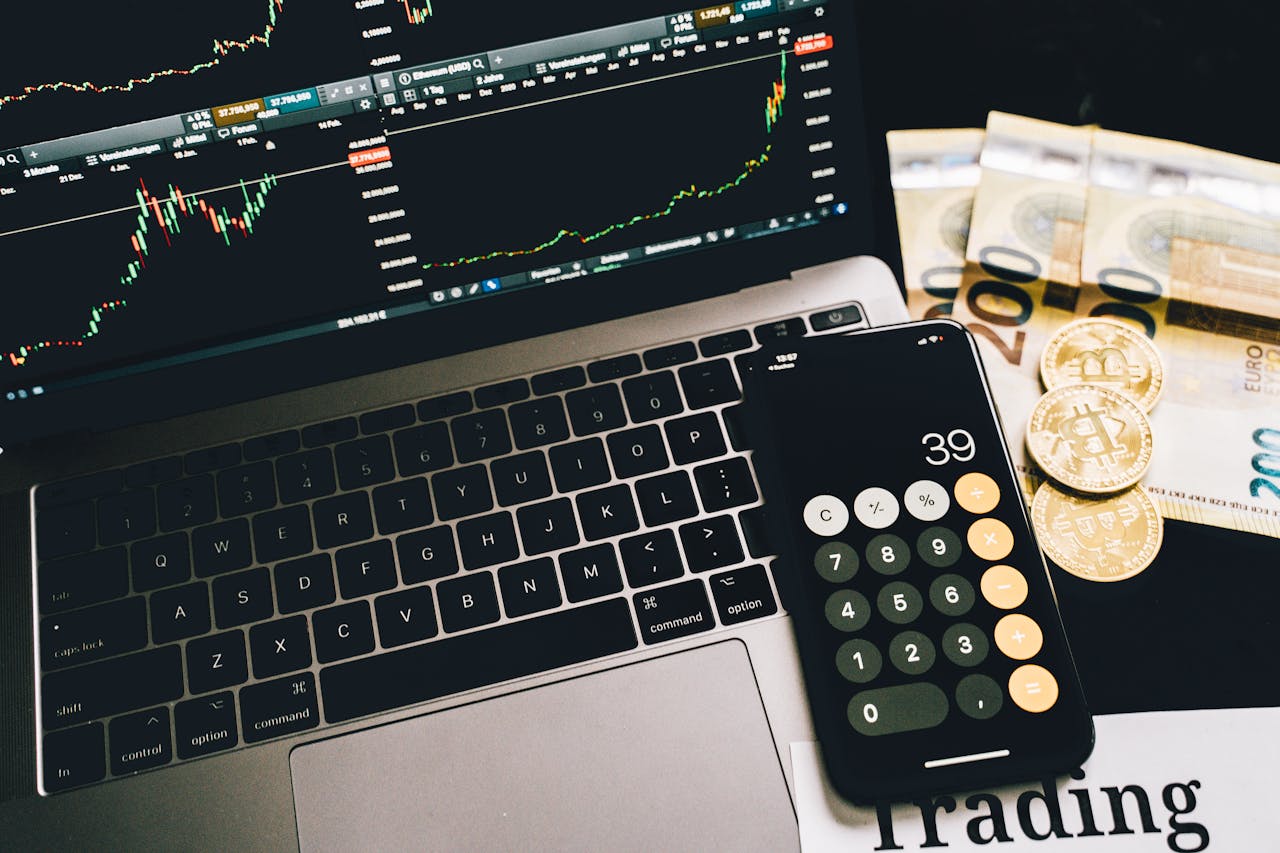 Laptop screen with trading charts, calculator app, Euros and Bitcoin coins on desk.